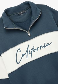 Sininen huppari vetoketjukauluksella, jossa on valkoinen vaakasuora raita ja "California"-teksti tummansinisellä skriptillä. Pehmeä kangas.