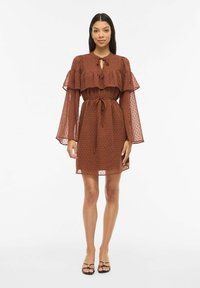 Robe marron à pois avec des manches volumineuses et transparentes à volants et une ceinture à nouer. Le tissu est léger et texturé, conçu pour une coupe décontractée.