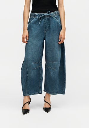 Femeie care poartă pantaloni largi scurți din denim albastru cu talie cu șnur și pantofi negri cu vârf ascuțit, pe fundal simplu.