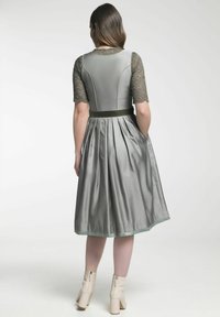 Grijze jurk met een aangesloten taille, kanten mouwen en een uitlopende rok. Heeft een gestructureerde stof en een groene satijnen riem. Draagt met crèmekleurige hakken.