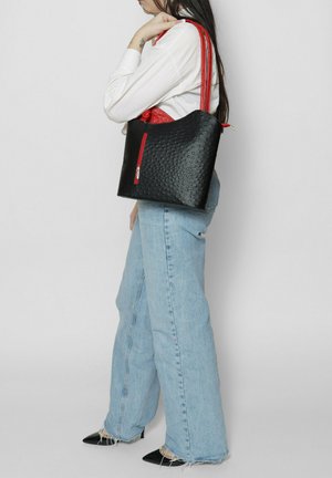 Schwarze Handtasche mit Struktur, rotem Reißverschluss und passenden Trägern, die ein geschwungenes Design aufweist. Wird an der Schulter zu hellen Jeans getragen.