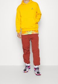 Žlutá bavlněná mikina s logem, vrstvená přes gradientový vzor. Hnědé nylonové kalhoty a vícebarevné sportovní boty doplňují outfit.