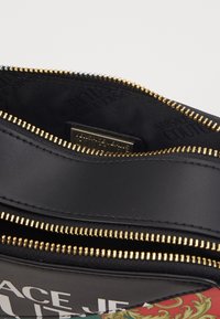 Sort lædertaske indvendig med guld lynlås, med et trykt design på ydersiden og et logo-tag synligt indeni.