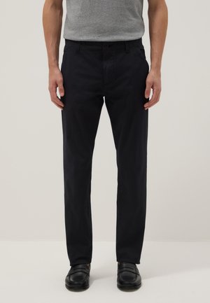 Hombre vistiendo pantalones negros de corte slim, camiseta gris metida por dentro y mocasines negros, de pie sobre un fondo claro y liso.