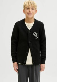 Juodas kardiganas su tekstūruotu mezginiu, turintis V-kaklą, penkis mygtukus ir siuvinėtą "QR" kairėje pusėje. Dėvimas virš šviesaus marškinėlio.