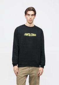 Mladý muž v černé mikině s žlutým logem „Santa Cruz Skateboards“, stojící před jednobarevným světlým pozadím s neutrálním výrazem.