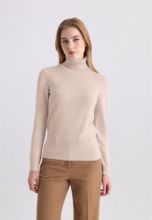 Une femme porte un pull à col roulé côtelé beige clair et un pantalon beige soutenu, sur un fond uni gris clair.