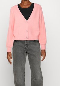 Rosa V-Ausschnitt-Strickjacke mit langen Ärmeln und Knopfverschluss, aus weichem Material gefertigt. Wird über einem schwarzen Top getragen, kombiniert mit grauen Jeans.