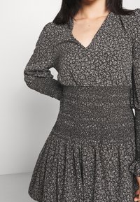 Černé květinové šaty s květinovým potiskem, s nařaseným pasem, dlouhými rukávy a rozšířenou sukní. Materiál má měkkou texturu.