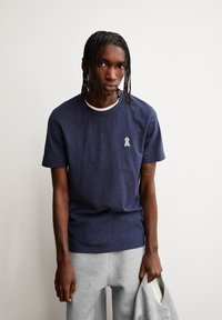 Marineblauw katoenen t-shirt met een ronde hals, met een klein grijs logo op de borst. G gedragen met lichtgrijze sweatpants.