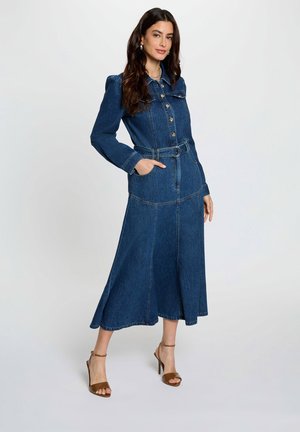 Vrouw met lang donker haar draagt een blauw denim midi-jurk met ceintuur en knopen, en bruine sandalen met hoge hakken, poseert met één hand in de zak.