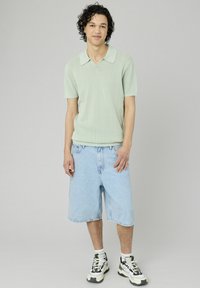 Polo shirt tricoté vert clair à manches courtes, associé à un short en denim bleu clair jusqu'aux genoux et à des baskets noir et blanc.