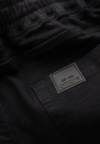 Shorts en denim noir avec une ceinture élastique, dotés d'une étiquette en cuir texturée portant l'inscription "ombire EST. 1985" sur la poche arrière.
