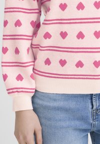 Pull rose pâle avec un motif de cœur en rose foncé, présentant des rayures horizontales et des poignets côtelés. Tissu en maille texturée.