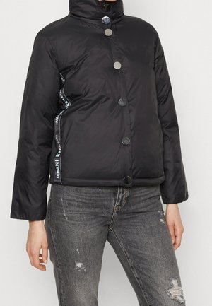 Sort dunjakke med trykknapper og mærket sidebånd båret med mørkegrå slidte jeans på en stående person.