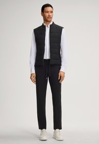 Zwarte gewatteerde vest met verticale stiksels, gedragen over een wit shirt. Gecombineerd met zwarte broek en witte sneakers. Eenvoudig en modern ontwerp.