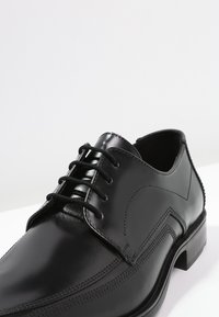 Chaussure habillée en cuir noir avec un bout arrondi, cinq œillets, texture lisse, couture décorative et un petit talon en bloc.