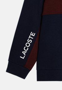 Felpa Lacoste con corpo di un blu scuro, accenti bordeaux, polsini a coste e testo del logo bianco sulla manica. Tessuto morbido.