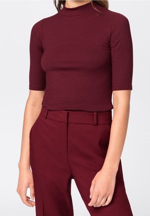 Femme portant un haut ajusté, côtelé, bordeaux à manches courtes, rentré dans un pantalon bordeaux taille haute avec des passants de ceinture.