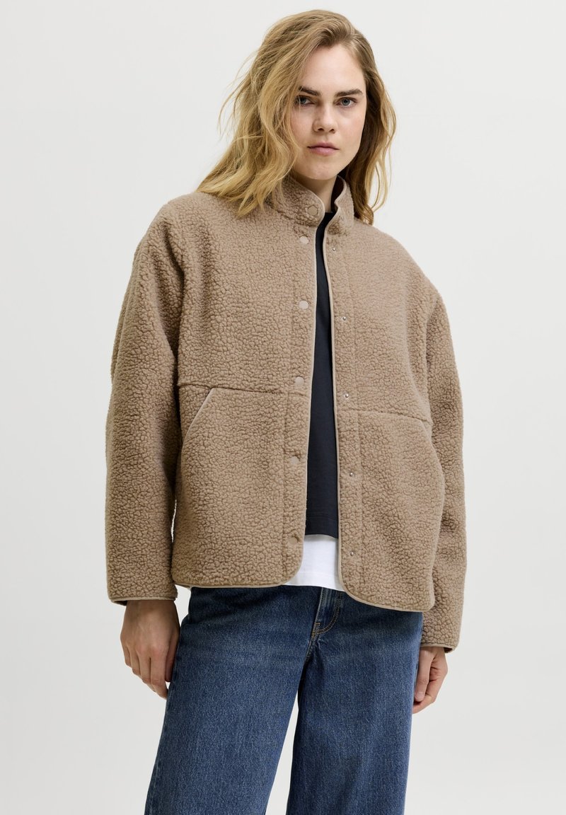 Γυναίκα με μπεζ σακάκι sherpa fleece με κουμπιά στερέωσης, μαύρο πουκάμισο, λευκό εσώρουχο και μπλε τζιν μπροστά από ένα απλό ανοιχτόχρωμο φόντο.