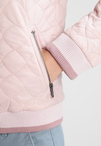 Hellrosa gesteppte Jacke mit silbernem Reißverschluss und Taschen, gerippten Bündchen und Saum mit dunkelpinkfarbenen Streifen, die Hand in der Tasche.