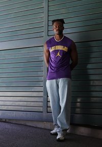 Tricou fără mâneci de culoare mov cu textul "LOS ANGELES", blugi albaștri deschis și adidași albi. Fundalul prezintă fâșii verzi orizontale.