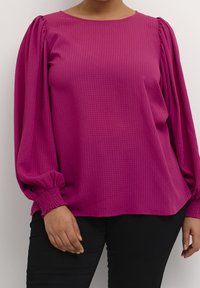 Femme portant un chemisier texturé magenta avec de longues manches bouffantes et des poignets smockés, associé à un pantalon noir, se tenant contre un mur blanc.
