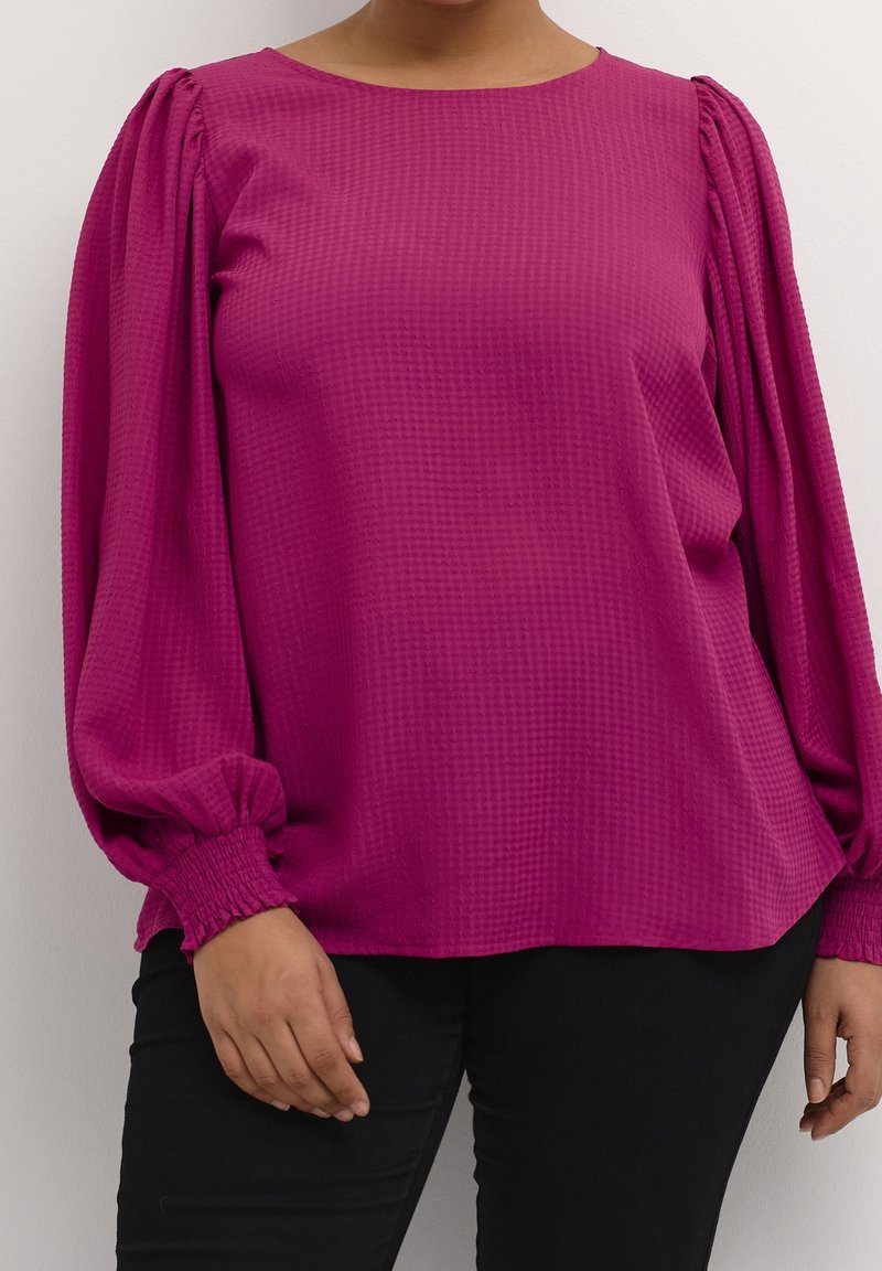 Femme portant un chemisier texturé magenta avec de longues manches bouffantes et des poignets smockés, associé à un pantalon noir, se tenant contre un mur blanc.