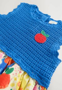 Haut en tricot bleu avec un bord festonné, présentant un appliqué en crochet de fruits rouges et verts. Motif floral coloré sur le tissu en dessous.