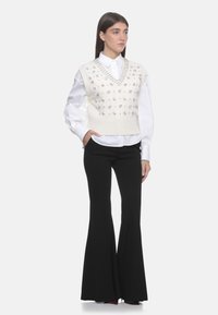 Gilet a V, in maglia color crema con decorazioni argentate, indossato sopra una camicia bianca. Abbinate a pantaloni neri a zampa d'elefante, caratterizzati da una texture liscia.