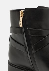 Bottine en cuir noir avec un design de sangle diagonale et une fermeture éclair dorée sur le côté. Texture lisse avec une forme structurée et sans motifs visibles.