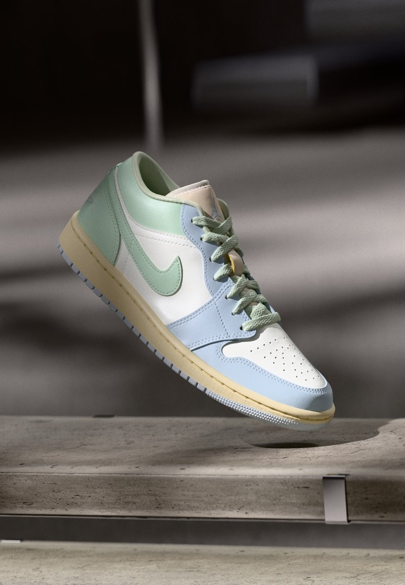 Basket basse en blanc, bleu pastel et vert menthe, présentée suspendue au-dessus d’un banc en béton sous un éclairage doux.