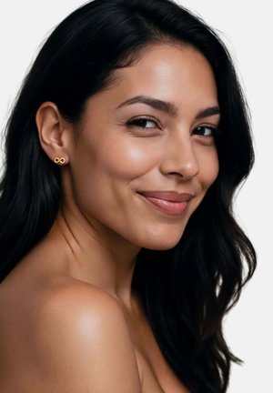 Tânără cu păr lung și închis la culoare, zâmbind subtil, purtând cercei mici cu simbolul infinitului din aur, pe un fundal simplu, deschis la culoare.