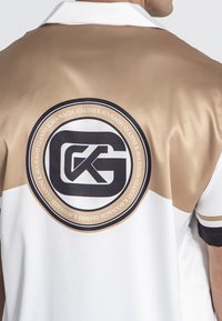 Camisa de manga corta en tela brillante beige y blanca, que presenta un prominente logo circular negro con las letras "GK" en la parte posterior.