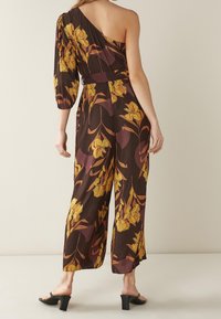 Jumpsuit de flores marrones con un hombro, que presenta flores amarillas y hojas verdes. Diseño de pierna ancha, tejido ligero y manga tres cuartos.