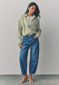 Camicia a righe verdi e bianche con bottoni, canottiera bianca, jeans di denim blu larghi, sandali beige con tacco. Il modello posa con una mano in tasca.