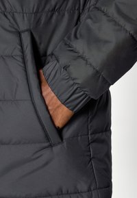 Μαύρο puffer μπουφάν με υφή καπιτονέ. Ορατή πλευρική τσέπη και ελαστική μανσέτα για άνετη εφαρμογή, με κοντινή λήψη του μανικ ιού και της τσέπης.