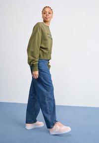 Maslinasto zeleni skraćeni dukserica, plavi traper široki ženski traperice i svijetlo ružičaste tenisice s bijelim potplatima. Opustan outfit, minimalni detalji.