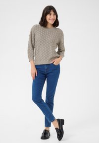 Beige gestrickter Pullover mit Rautenmuster, Dreiviertelärmeln und rundem Ausschnitt, kombiniert mit blauen Jeans und schwarzen Mokassins.