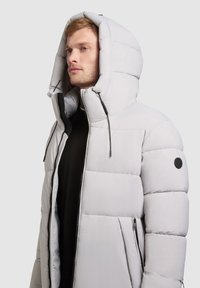 Hellgraue Pufferjacke mit großer Kapuze, Reißverschluss vorne und seitlichen Taschen. Sie hat ein gestepptes Design und schwarze Akzente an den Ärmeln.