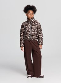 Piumino con stampa leopardata, collo alto, cerniera nera e polsini a costine, abbinato a pantaloni marroni a gamba larga e sneakers a righe.