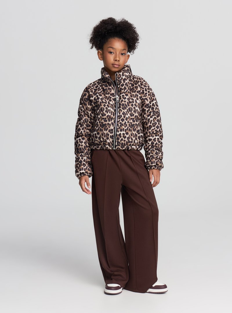 Piumino con stampa leopardata, collo alto, cerniera nera e polsini a costine, abbinato a pantaloni marroni a gamba larga e sneakers a righe.