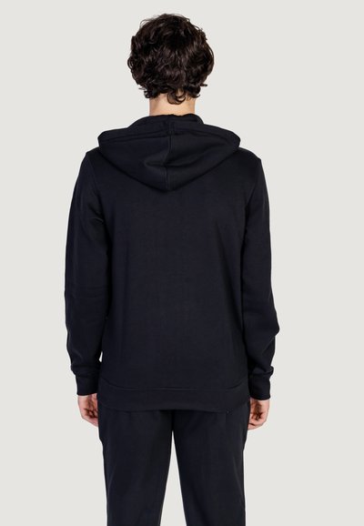 Haut à capuche en coton noir avec un cordon de serrage, texture lisse, poignets et ourlet côtelés ; porté avec un pantalon assorti, vu de l'arrière.