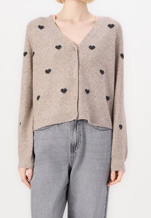 Cardigan beige boutonné avec des motifs de cœurs noirs porté sur un top rose clair, associé à un jean gris taille haute.