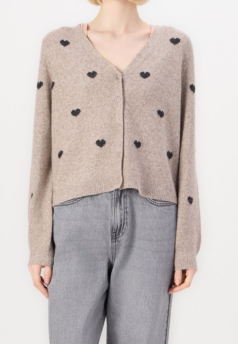 Cardigan beige boutonné avec des motifs de cœurs noirs porté sur un top rose clair, associé à un jean gris taille haute.