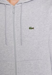 Sudadera ligera de color gris claro con cremallera, cordones y un pequeño logo de cocodrilo verde bordado en el pecho.