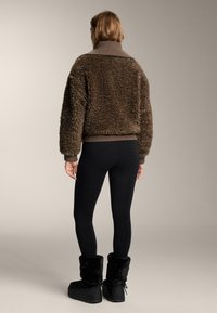 Bruin, zacht, kort jasje met een hoge kraag en geribbelde zoom, gecombineerd met zwarte legging en zwarte laarzen met bontvoering. Gladde, zachte textuur.