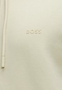 Tissu couleur crème avec texte « BOSS » en relief et cordon assorti avec embout en plastique sur le côté gauche.