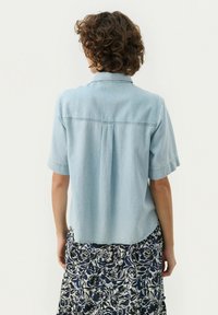 Light blue short-sleeve shirt made of soft fabric, featuring a collar, back pleat detail, and a curved hemline. Patterned skirt below.