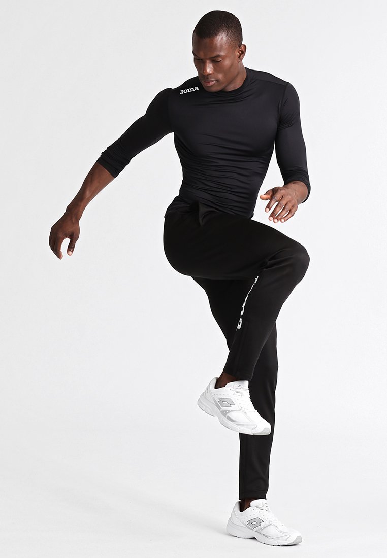 Zwarte sportshirt met lange mouwen en een aansluitend ontwerp, met een wit logo, gecombineerd met zwarte smalle broek en witte sneakers.
